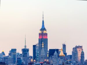 The image shows the New York City skyline at dusk, with the Empire State Building glowing in red, white, and blue. Silhouetted against a pastel sky stand skyscrapers of varying architectural styles, much like how the city's most visited museums reflect a blend of historic and modern influences.